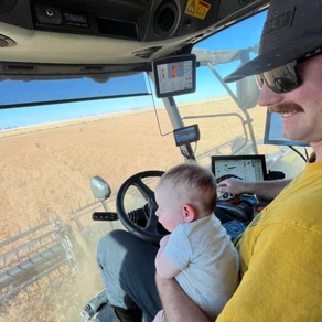 A baby riding in the cab of a combine, held by his father, during harvest